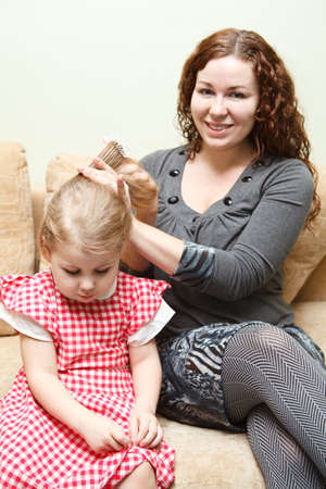 Mother and little daughter sitting on couch. Woman combing daughter hairの写真素材
