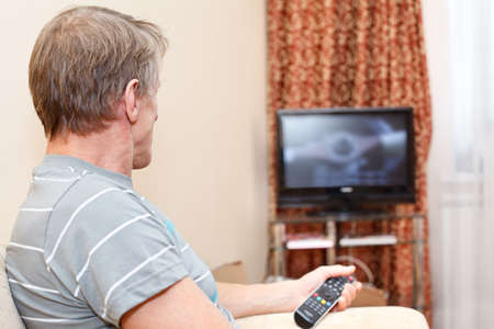 Senior man with remote control sitting on couch and looking at tv set