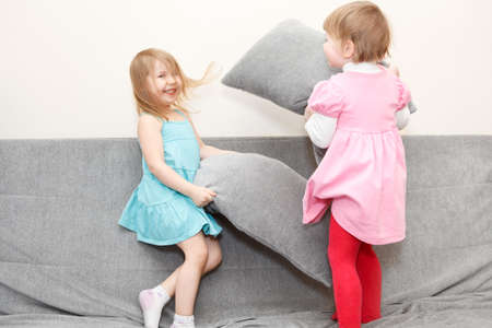 Two Caucasian happy playful little girls in pillows fighting on sofaの写真素材
