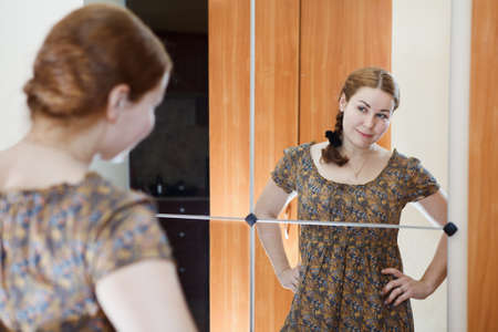 Portrait of young Caucasian female in dress standing against mirror and looking at herself in domestic roomの写真素材