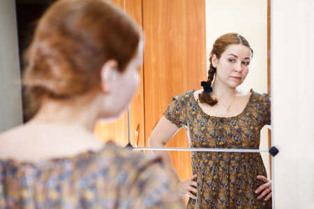 Portrait of young Caucasian female in dress standing against mirror and looking at herself in domestic roomの写真素材