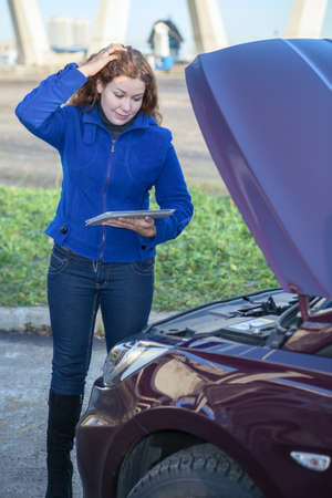 Woman in thought standing before car with tablet pc and opened hoodの写真素材