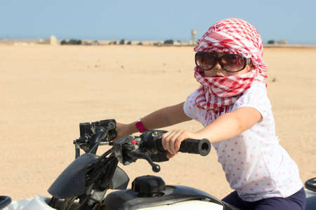 Small Caucasian child in head kerchief and sunglasses on quadbike in desertの写真素材