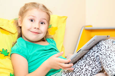 Little Caucasian girl with funny smile and wide opened eyes sitting with tablet computerの写真素材