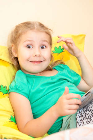 Little caucasian girl with wide open eyes sitting with tablet computer in hands in bedの写真素材
