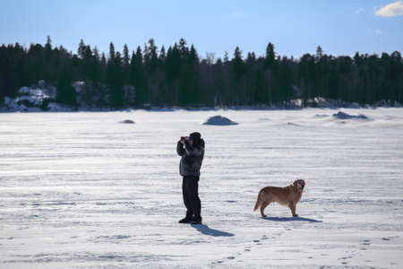 Man with dog looking at binocular and standing on frozen lakeの写真素材