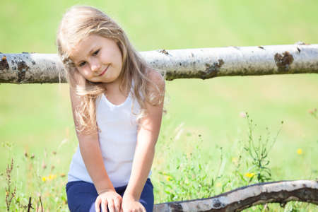 Embarrassed Caucasian small girl sitting on rural fence. Copyspaceの写真素材