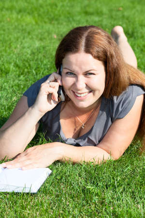 Happy smiling young woman with book speaking by phone on green grassの写真素材