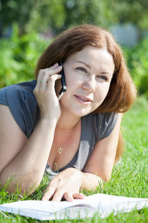 Beauty Caucasian woman calling on the phone and reading the book on green meadowの写真素材