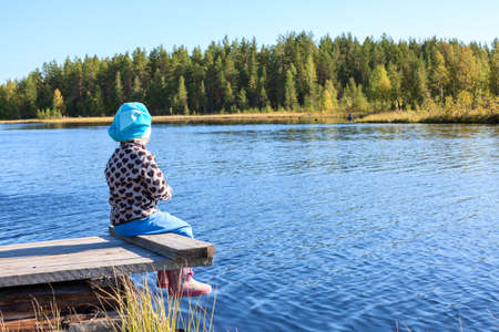 Little child sitting on wooden jetty lake front, legs dangling in blue water, copyspaceの写真素材