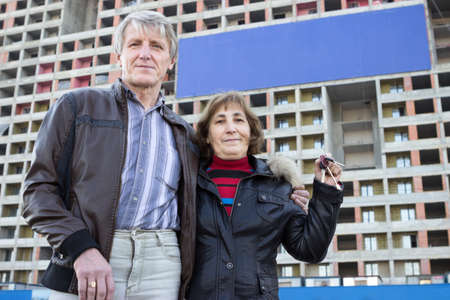 Senior couple standing with house keys in hand against blue blank banner on buildingの写真素材