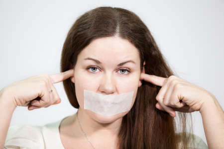 Caucasian young woman with mouth sealed and plugged ears, close-up, grey backgroundの写真素材