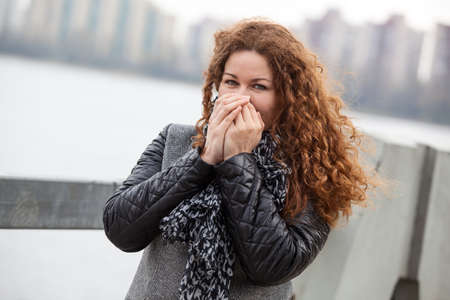Frozen Caucasian woman with long curly hair warming her hands while standing in the wind on the city streetの写真素材