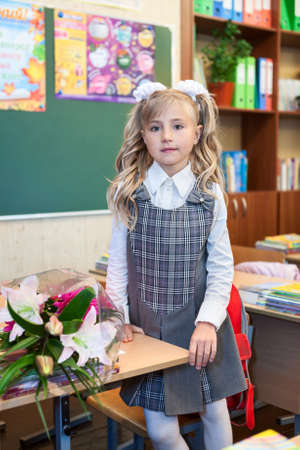 Cute schoolgirl in school uniform standing near desk in classroom