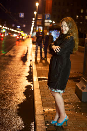 Pretty frozen woman in black coat and high heels walking alone on the pavement in the night cityの写真素材
