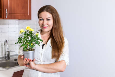Young woman housewife stands at kitchen, holding yellow roses at pot in hands, copy spaceの写真素材