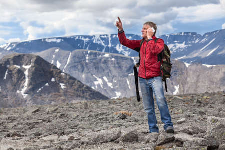 Mature Caucasian hiker a man shows to the mountain top, talking on a cell phoneの写真素材