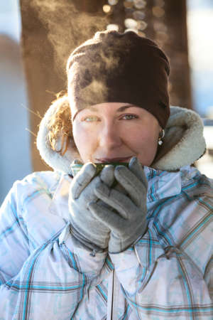 Young woman drinking hot tea from mug holding it in hands with mittens, winter season, outdoorsの写真素材