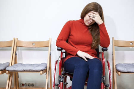 Paralyzed legs sad woman in invalid chair covers her face with hand while sitting indoorsの写真素材