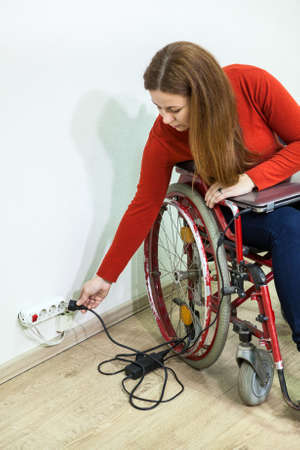 Young disabled Caucasian woman sitting wheelchair with power plug in hand, stretching to power socket in wallの写真素材