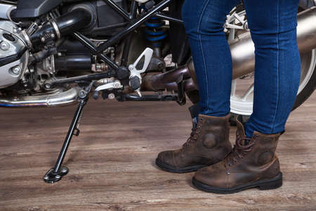 Female legs wearing brown leather safety boots for motorcycling are near motorbike, close up view