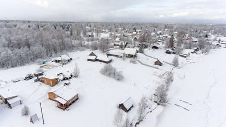 Rural timber houses and barns are on winter lake shore. Russian remote village in evergreen forests. Aerial view. Russiaの写真素材