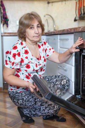 Mature Caucasian woman sitting beside the built-in oven with open door. Domestic kitchenの写真素材