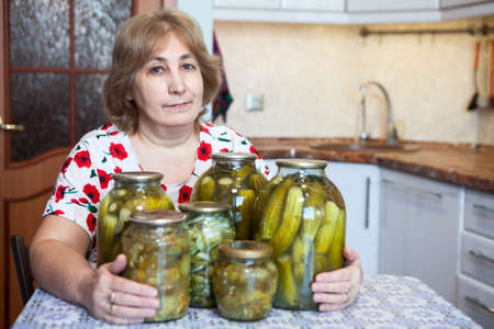 Caucasian Russian middle-aged woman sits at a table in kitchen with glass jars full of picklesの写真素材