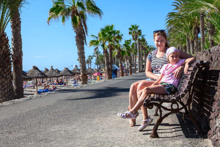 Woman and young girl sitting together on the bench in palm tree alley near the beach. Copy space. Tenerife, Canaryの写真素材