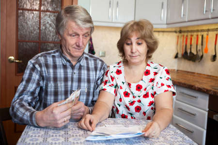 Surprised husband and wife looking at the bills with cash money in hands, domestic kitchenの写真素材