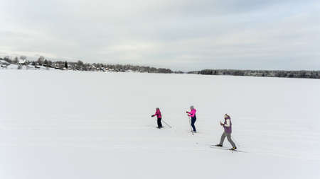Three people skiing in frozen snowy lake to the village on shore, winter in Russiaの写真素材