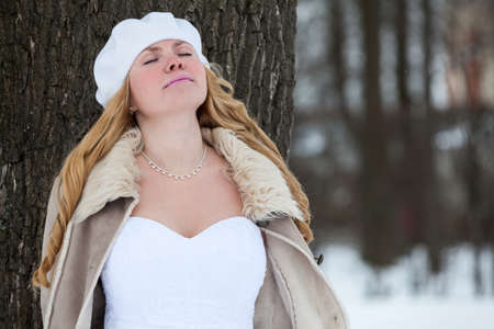 Young Caucasian bride in white wedding dress and fur coat leaning to tree trunk with closed eyesの写真素材
