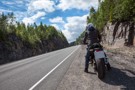 Motorbike driver is ready to start moving on the asphalt route, sitting on motorbike on roadside, rear viewの写真素材