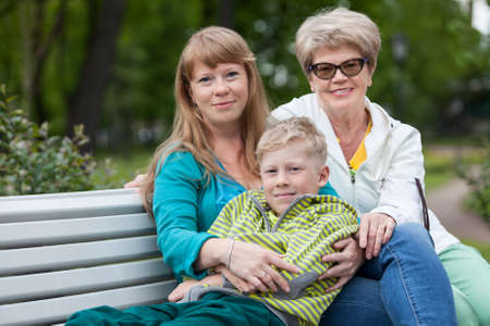 Family of three people looking at camera sitting on garden bench together in hugs, young boy, adult...