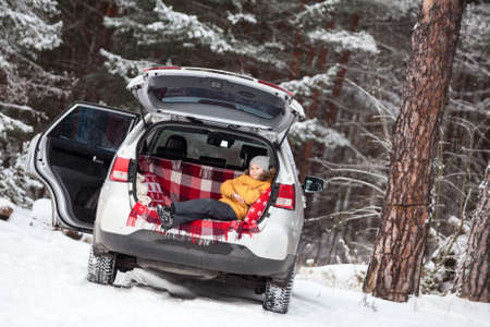 Smiling child laying inside of car back boot for luggage. Winter evergreen forest with huge snow...