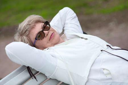An elderly woman in glasses rests on her back, on benchの写真素材