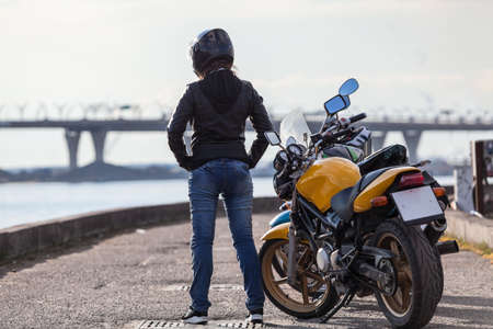 Rear view at girl biker in motorcycle helmet standing next to several bikes on street sea embankment, full-lengthの写真素材