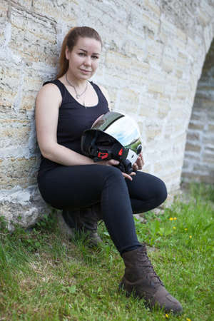 Caucasian woman with crash helmet in hands and protective boots sitting on green grassの写真素材