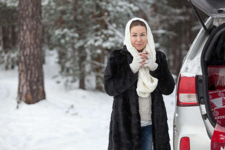 Young European woman standing near her car and holding tea mug in hands at winter season, copy spaceの写真素材