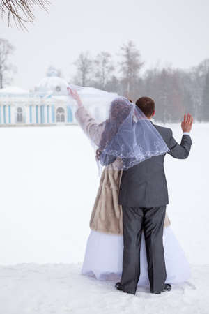 Russian newlywed couple standing backside near frozen lake and embracing. Blizzard and cold weather, winter timeの写真素材