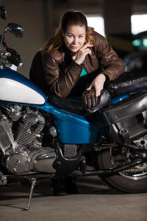Biker woman with heavy build standing near her motorcycle, leaning the seat, looking at cameraの写真素材