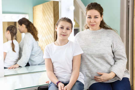 Portrait of adult mother and her teenager daughter sitting in dressing room, looking at camera
