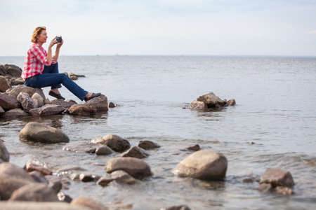 Caucasian woman sitting on stones near the water. Making pictures with cellphone, calm seaの写真素材
