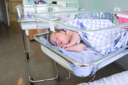 Cute little newborn baby sleeping in infant bed in hospital, folded hands with open mouth, under blanketの写真素材