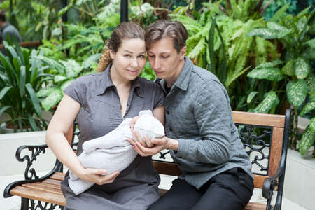 Portrait of happy parents with newborn baby in hands, sitting on bench in green plants
