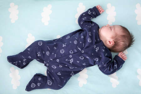 One month old infant sleeping in bed with hands up, dark blue clothes, light blue linen with clouds, full length viewの写真素材