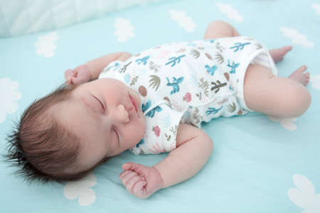 Sleeping infant dressed romper suit, baby lying on bedsheet with closed eyes, hands up