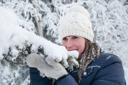 Portrait of Caucasian smiling woman touching snow cap on fir tree branch with her mittens, winter...
