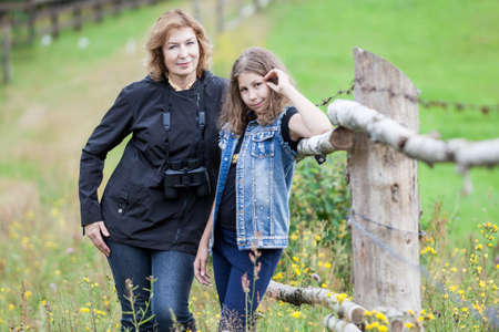 Portrait of mature mother and young daughter standing together near farm paddock, looking at camera,...