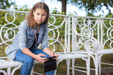 Thoughtful teen age girl sitting on bench with smartphone in hands, looking far away, copyspaceの写真素材